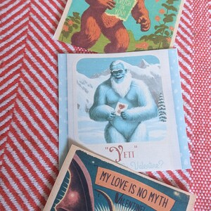 May include: Three Valentine's Day cards featuring illustrations of a Bigfoot, Yeti, and a Mothman. The Bigfoot card says "I have a "BIG" crush on you!" The Yeti card says "Yeti be my Valentine?" The Mothman card says "My love is no myth Valentine!"