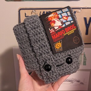 May include: A gray crocheted pouch featuring two black plastic bead eyes and a stitched smile. A rectangular tag is attached to the front, displaying the Super Mario Bros. Entertainment System game cover art with the character Mario in pixelated form.