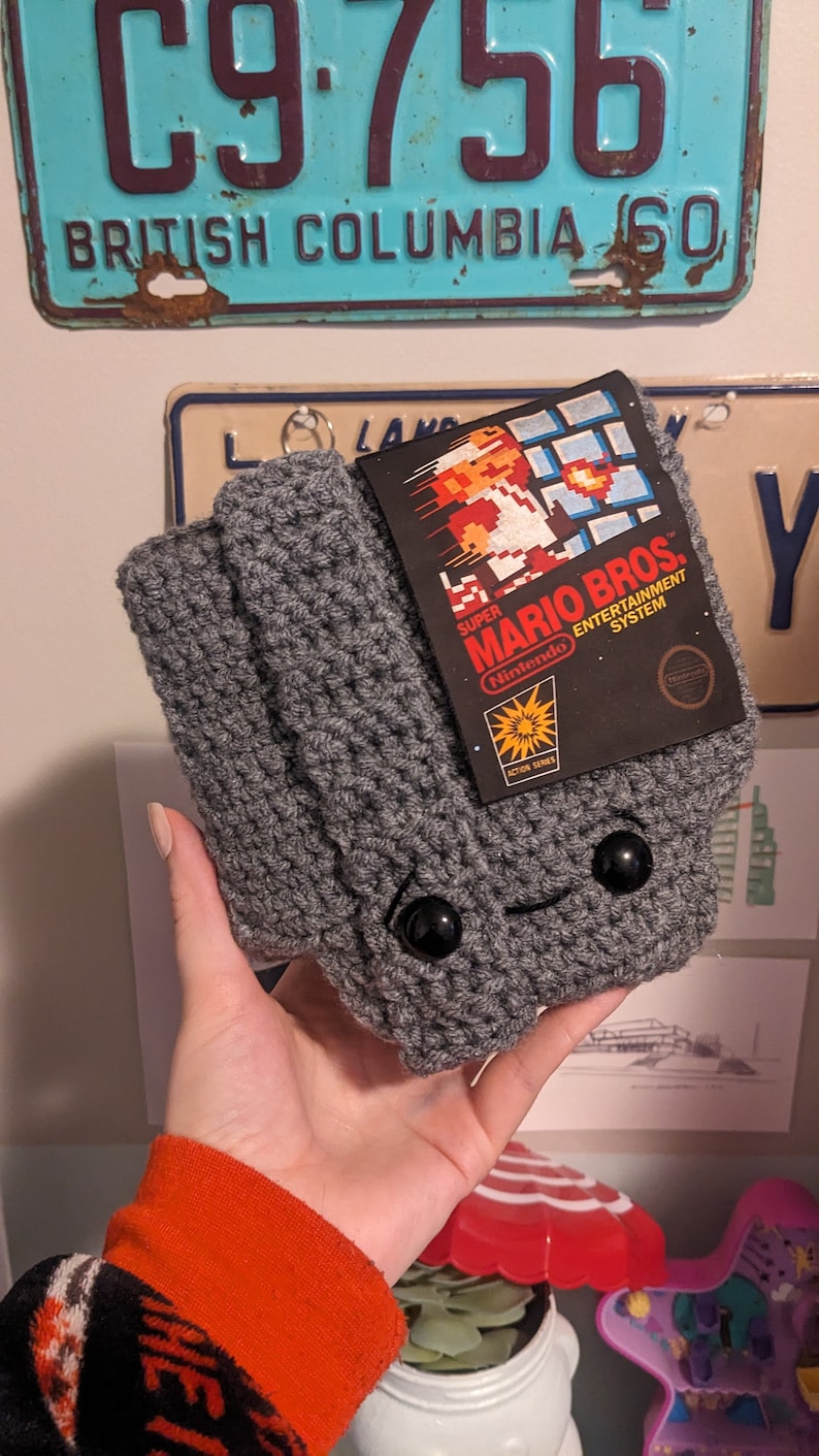 May include: A gray crocheted pouch featuring two black plastic bead eyes and a stitched smile. A rectangular tag is attached to the front, displaying the Super Mario Bros. Entertainment System game cover art with the character Mario in pixelated form.