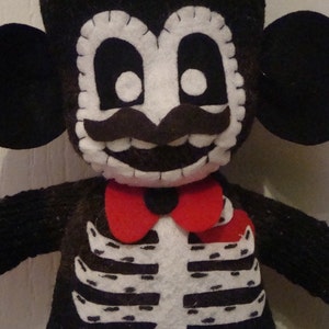 May include: A black and white felt toy monkey dressed as a skeleton with a red bow tie. The monkey has a white face with black eyes, a black mustache, and a toothy grin.