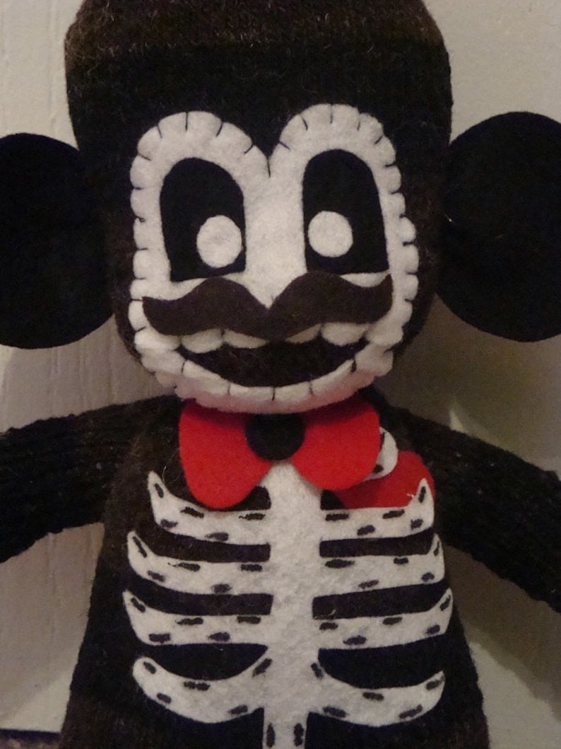 May include: A black and white felt toy monkey dressed as a skeleton with a red bow tie. The monkey has a white face with black eyes, a black mustache, and a toothy grin.