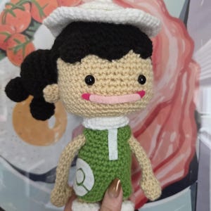 May include: A handmade crochet doll with a white hat, black hair, and a green outfit. The doll has a light skin tone, black eyes, and a pink mouth. The doll's shoes are yellow and white.