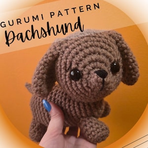 Op de afbeelding: Een gehaakte bruine teckel amigurumi. De handgemaakte hond heeft grote zwarte ogen en een zwarte neus. De afbeelding bevat de tekst "AMIGURUMI PATTERN Dachshund" in zwart script. De achtergrond is oranje met een witte en oranje gradiënt.