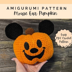 Puede incluir: Una calabaza de ganchillo naranja con un tallo marrón y dos orejas negras redondas. La calabaza presenta una cara de fieltro negro con ojos ovalados, una nariz redonda y una boca ancha sonriente. Texto: AMIGURUMI PATTERN Mouse Ear Pumpkin y Easy PDF Crochet Pattern.