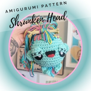 May include: A crocheted amigurumi toy in the shape of a shrunken head with a rainbow colored yarn hair. The head has a happy expression with a red mouth and black eyes with an X over each eye. The toy is hanging from a wooden stick.
