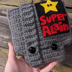 May include: A gray crocheted hat featuring a black rectangular patch with a yellow star and the red text "SUPER ALAIN" stitched on the front. The hat includes two black button eyes and a ribbed cuff design, crafted with a textured yarn finish.