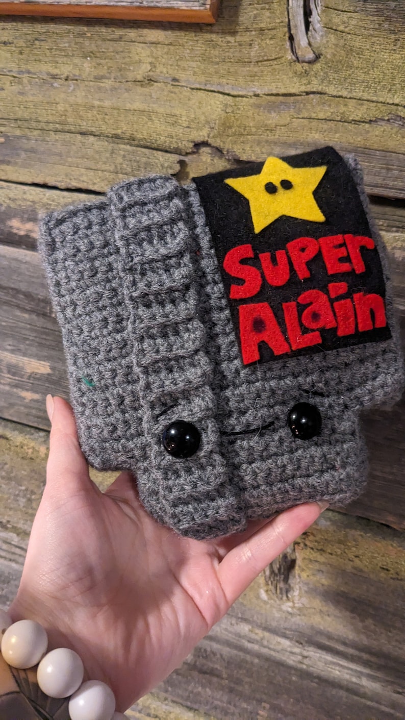May include: A gray crocheted hat featuring a black rectangular patch with a yellow star and the red text "SUPER ALAIN" stitched on the front. The hat includes two black button eyes and a ribbed cuff design, crafted with a textured yarn finish.