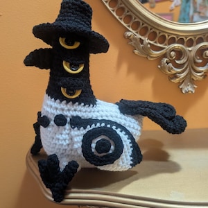 Pode incluir: Uma criatura de crochê feita à mão com um chapéu preto e três olhos amarelos. O corpo é branco com detalhes em preto, incluindo asas e padrões circulares. O design caprichoso é exibido em uma prateleira decorativa.