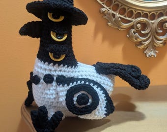 Peut inclure: Une créature au crochet faite à la main avec un chapeau noir et trois yeux jaunes. Le corps est blanc avec des accents noirs, y compris des ailes et des motifs circulaires. Le design fantaisiste est exposé sur une étagère décorative.
