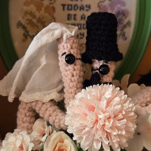 Puede incluir: Una pareja de bodas de ganchillo caprichosa con ojos de botón negros, velo y sombrero de copa, rodeada de flores rosas y blancas. El fondo presenta un punto de cruz enmarcado con el texto "LET TODAY BE A..."