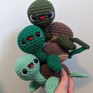 May include: A stack of three crocheted green and brown turtle toys with smiling faces. The turtles are stacked on top of each other, with the largest turtle on the bottom and the smallest turtle on top.