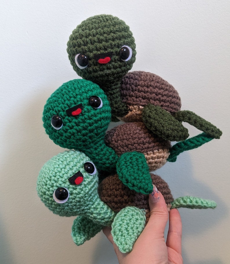 May include: A stack of three crocheted green and brown turtle toys with smiling faces. The turtles are stacked on top of each other, with the largest turtle on the bottom and the smallest turtle on top.
