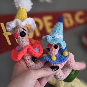 May include: Two handmade crochet figures, each with a whimsical design. One wears a yellow hat with a white pom-pom and a red and orange collar. The other has a blue hat and collar. Both have black eyes and red noses.