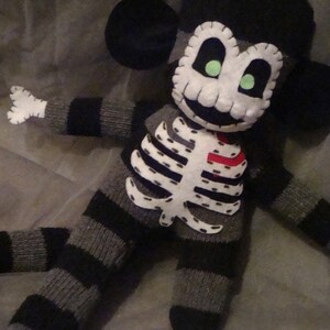 May include: A black and gray striped sock monkey with a white felt skull and crossbones design. The monkey has green felt eyes and a red felt heart on its chest.