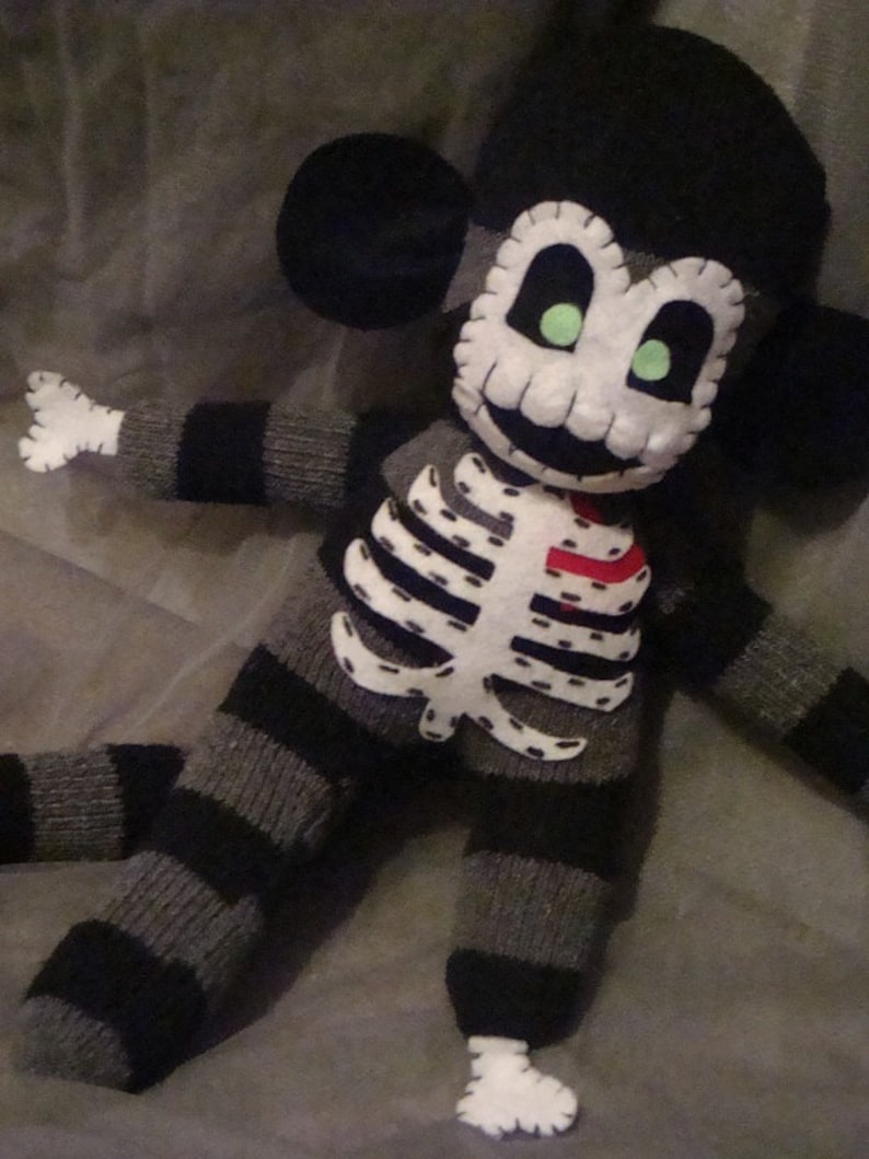 May include: A black and gray striped sock monkey with a white felt skull and crossbones design. The monkey has green felt eyes and a red felt heart on its chest.