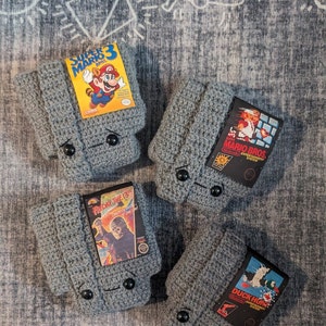 May include: Four grey crocheted covers designed to resemble Nintendo Entertainment System game cartridges. Each cover features a black button closure and a rectangular label depicting Super Mario Bros. 3, Super Mario Bros., Friday the 13th, and Duck Hunt.