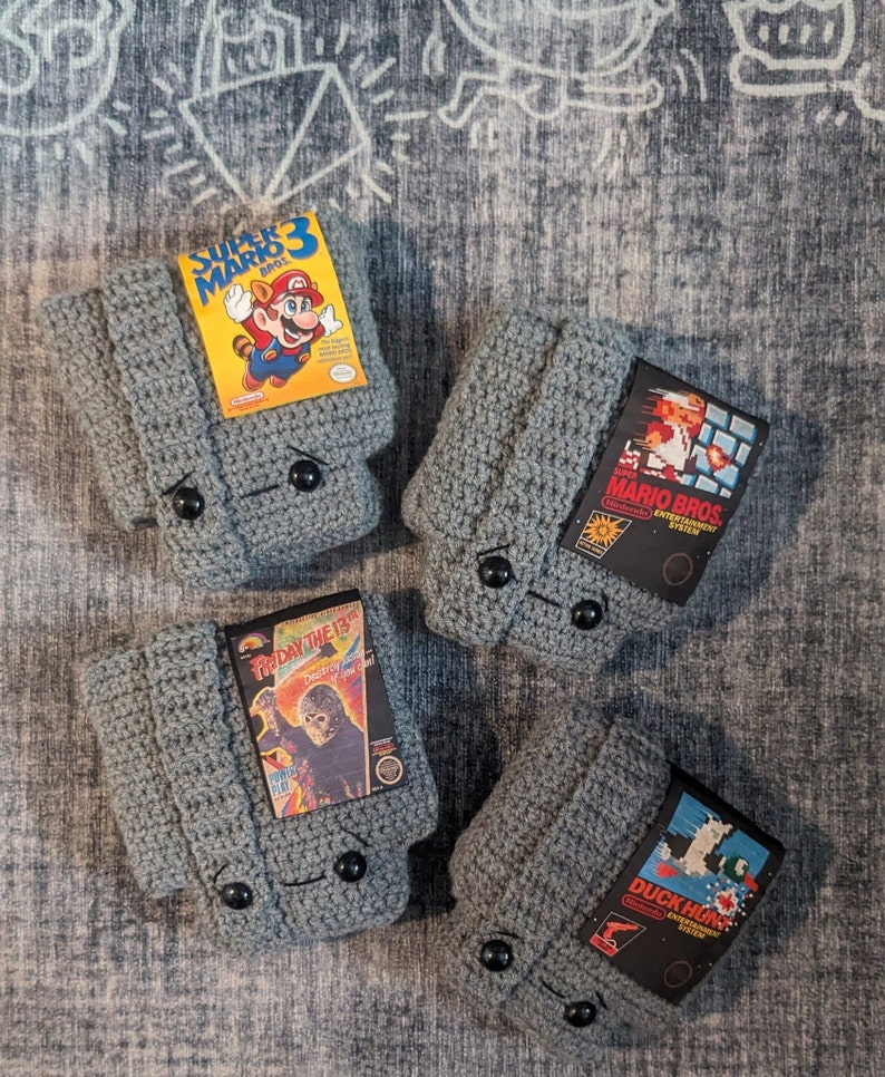 May include: Four grey crocheted covers designed to resemble Nintendo Entertainment System game cartridges. Each cover features a black button closure and a rectangular label depicting Super Mario Bros. 3, Super Mario Bros., Friday the 13th, and Duck Hunt.