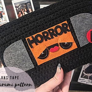 Puede incluir: Un patrón amigurumi de cinta VHS negra tejida con un fieltro naranja que dice "HORROR". La etiqueta tiene ojos de botón y una "R" roja de fieltro. También se ve el texto "VHS TAPE amigurumi pattern".