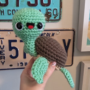 May include: A crocheted green turtle with a brown shell and a happy face. The turtle is being held by a hand.