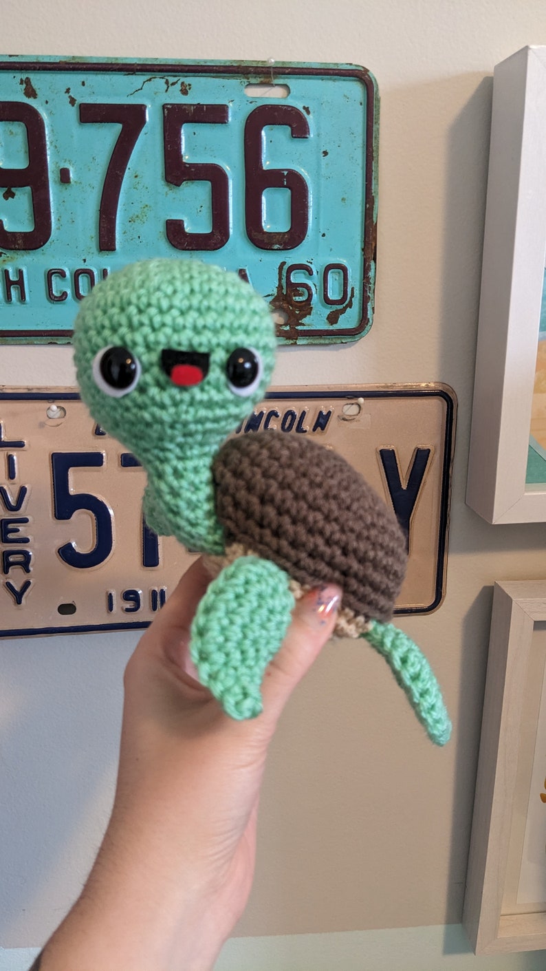 May include: A crocheted green turtle with a brown shell and a happy face. The turtle is being held by a hand.