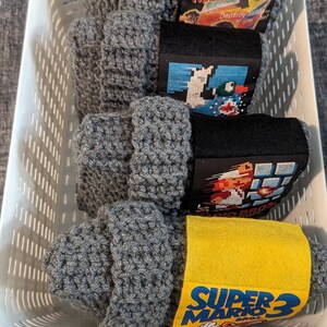 May include: Four gray crocheted beanies stacked in a white plastic basket. Each beanie features a paper band with retro video game art, including Friday the 13th, Duck Hunt, Super Mario Bros, and Super Mario Bros 3, with clear text labels.