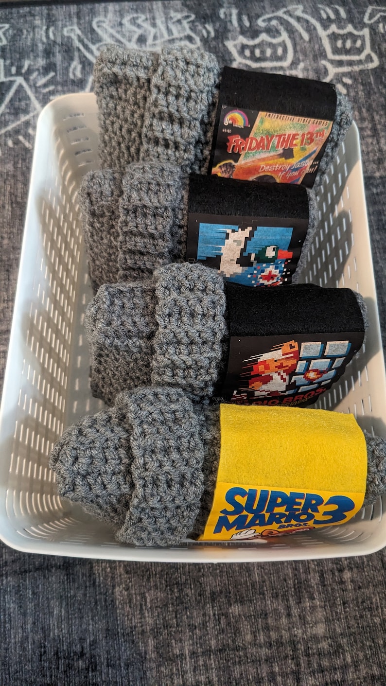 May include: Four gray crocheted beanies stacked in a white plastic basket. Each beanie features a paper band with retro video game art, including Friday the 13th, Duck Hunt, Super Mario Bros, and Super Mario Bros 3, with clear text labels.