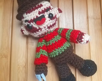 May include: A crocheted plush toy of Freddy Krueger, the villain from the Nightmare on Elm Street movie franchise. The toy is wearing a red and green striped sweater, a brown hat, and has a brown face with black eyes and a red mouth. The toy is holding a silver knife in its right hand.