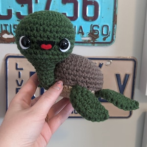 May include: A crocheted green sea turtle with brown shell and black eyes. The turtle has a red mouth and is being held by a hand.