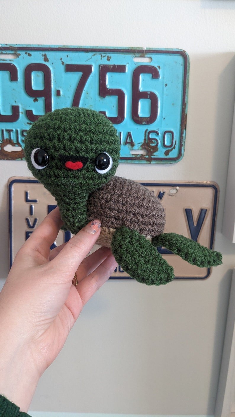 May include: A crocheted green sea turtle with brown shell and black eyes. The turtle has a red mouth and is being held by a hand.