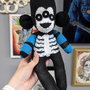 May include: A black sock monkey doll with a white skeleton design and blue and black striped arms. The doll has three large black buttons for eyes and a stitched-on smile.