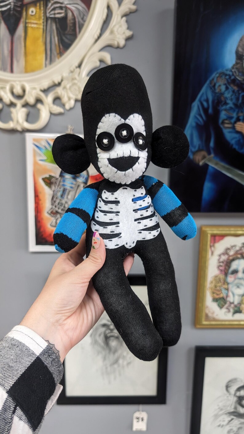 May include: A black sock monkey doll with a white skeleton design and blue and black striped arms. The doll has three large black buttons for eyes and a stitched-on smile.