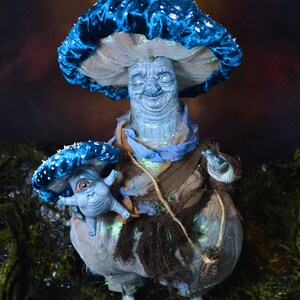 Puede incluir: Una figurita en forma de hongo azul y blanco con una cara sonriente, sosteniendo una figurita m&aacute;s peque&ntilde;a en forma de hongo azul y blanco con un ojo grande. Ambas figuritas est&aacute;n hechas de un material texturizado y tienen una est&eacute;tica caprichosa y de cuento de hadas.