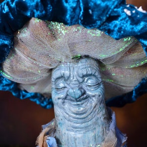Puede incluir: Un personaje de hongo azul y blanco con una cara sonriente. El hongo tiene un sombrero de terciopelo azul con detalles blancos y un velo de malla blanca.