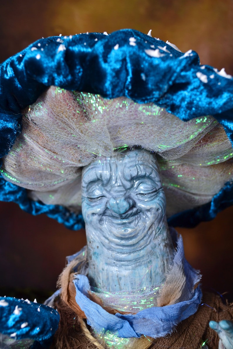 Puede incluir: Un personaje de hongo azul y blanco con una cara sonriente. El hongo tiene un sombrero de terciopelo azul con detalles blancos y un velo de malla blanca.