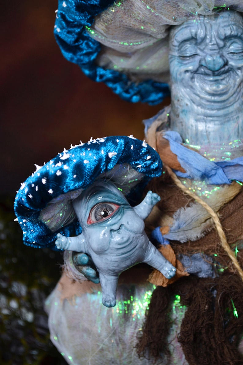 Puede incluir: Una mu&ntilde;eca con forma de hongo azul y blanco con un solo ojo grande y una cara sonriente. La mu&ntilde;eca est&aacute; siendo sostenida por una mu&ntilde;eca m&aacute;s grande de color azul y blanco con un sombrero blanco y una larga barba blanca.