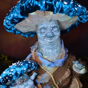 Puede incluir: Una figurita en forma de hongo azul y blanco con una cara sonriente y una figurita de hongo m&aacute;s peque&ntilde;a con un ojo. Ambas figuritas est&aacute;n hechas de arcilla y tienen una est&eacute;tica caprichosa de cuento de hadas.