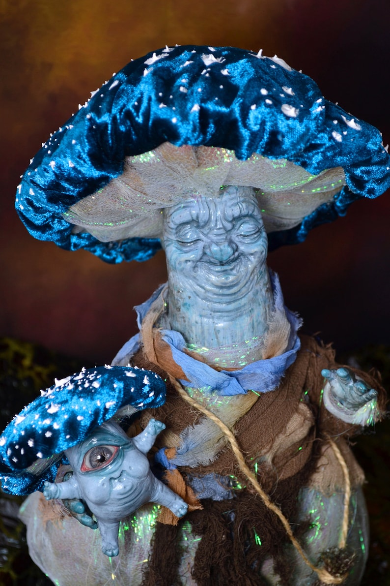 Puede incluir: Una figurita en forma de hongo azul y blanco con una cara sonriente y una figurita de hongo m&aacute;s peque&ntilde;a con un ojo. Ambas figuritas est&aacute;n hechas de arcilla y tienen una est&eacute;tica caprichosa de cuento de hadas.