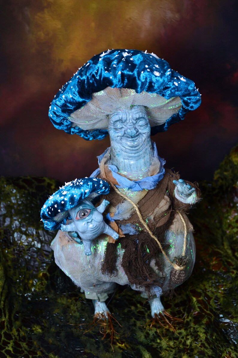 Puede incluir: Una figurita en forma de hongo azul y blanco con una cara sonriente y una figurita de hongo m&aacute;s peque&ntilde;a con un ojo en su mano.