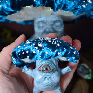 Puede incluir: Una figurita en forma de hongo azul y blanco con un solo ojo grande y una expresi&oacute;n caprichosa. La figurita est&aacute; hecha de un material texturizado y tiene un sombrero de terciopelo azul con manchas blancas.