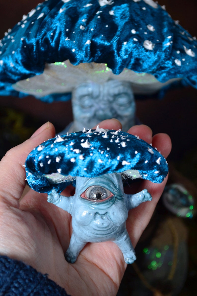 Puede incluir: Una figurita en forma de hongo azul y blanco con un solo ojo grande y una expresi&oacute;n caprichosa. La figurita est&aacute; hecha de un material texturizado y tiene un sombrero de terciopelo azul con manchas blancas.
