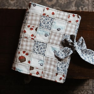 May include: A quilted journal cover with a patchwork design featuring strawberries, floral patterns, and gingham squares. A matching fabric bow secures the cover. The cover is resting on a dark wooden surface.