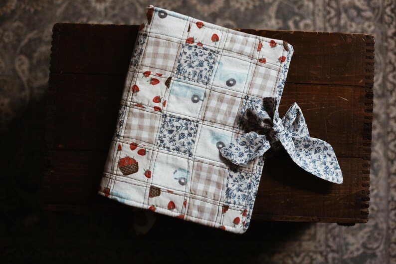 May include: A quilted journal cover with a patchwork design featuring strawberries, floral patterns, and gingham squares. A matching fabric bow secures the cover. The cover is resting on a dark wooden surface.
