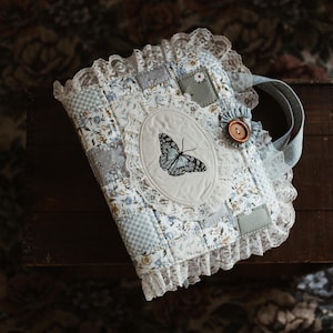 May include: A fabric-covered journal featuring a patchwork design in shades of blue, white, and grey. The cover displays a central oval panel with a black butterfly illustration, surrounded by white lace trim, a brown button, and fabric ruffles.
