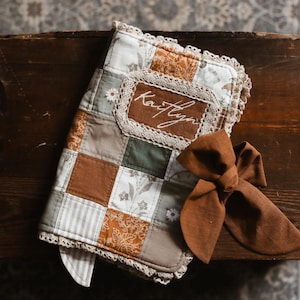 May include: A patchwork baby blanket with a brown bow. The blanket has squares of brown, green, and white fabrics, with lace trim. The name "Kaitlyn" is embroidered on a brown patch. The blanket is likely made of cotton.