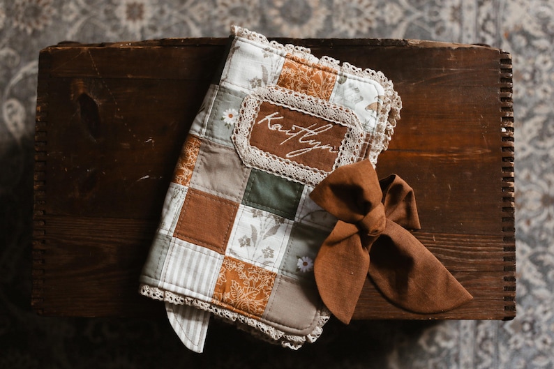 May include: A patchwork baby blanket with a brown bow. The blanket has squares of brown, green, and white fabrics, with lace trim. The name "Kaitlyn" is embroidered on a brown patch. The blanket is likely made of cotton.