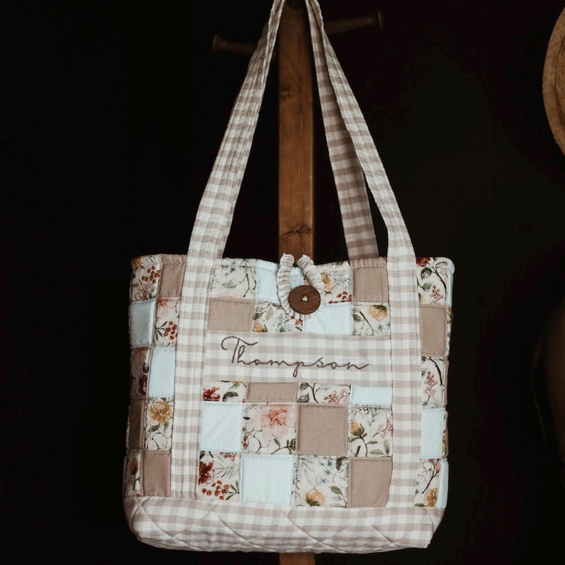 May include: A quilted tote bag with a patchwork design. The bag features squares of floral fabric, solid beige, and light blue, framed by a beige and white gingham pattern. The name "Thompson" is embroidered on the front, and a wooden button secures the top.