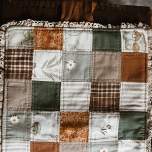May include: A patchwork quilt square in shades of brown, green, and cream. The design features floral and plaid patterns, with a delicate lace trim. The quilt is likely handmade. The measurements are in centimetres.