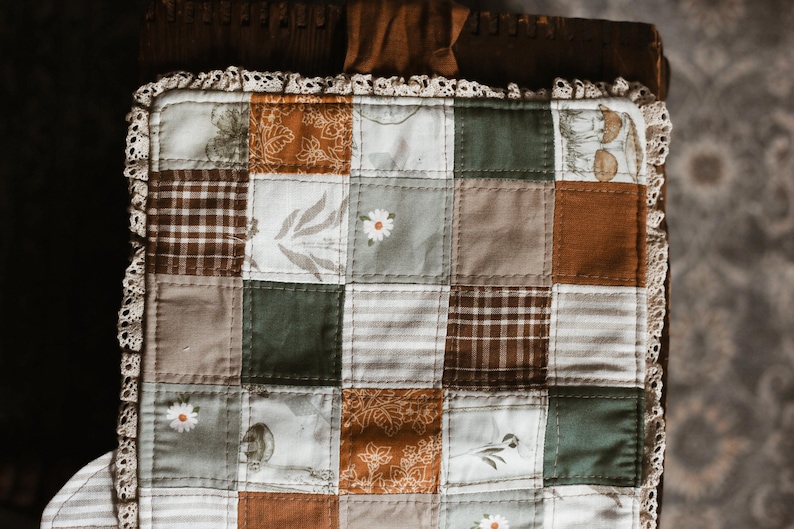 May include: A patchwork quilt square in shades of brown, green, and cream. The design features floral and plaid patterns, with a delicate lace trim. The quilt is likely handmade. The measurements are in centimetres.