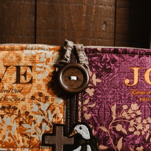 May include: Close-up of a fabric item with the word "LOVE" and a cross. The item features a duck applique and a large wooden button. The fabric has floral and paisley patterns in orange, purple, and green.