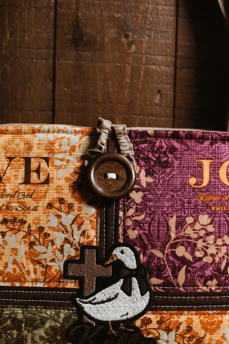 May include: Close-up of a fabric item with the word "LOVE" and a cross. The item features a duck applique and a large wooden button. The fabric has floral and paisley patterns in orange, purple, and green.